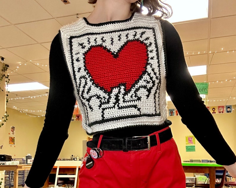 May include: A white crocheted vest with a black border and a red heart design. The vest is worn over a black long-sleeved shirt and red pants.