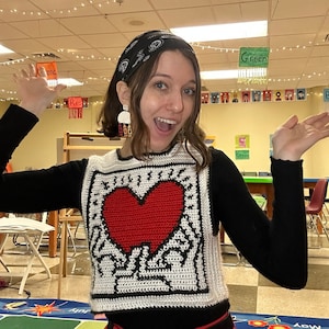 May include: A woman wearing a black sweater with a white crocheted heart design. The heart is red and has a black outline. The woman is wearing a black bandana on her head and has a bright smile.