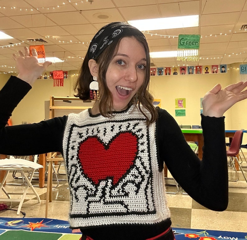 May include: A woman wearing a black sweater with a white crocheted heart design. The heart is red and has a black outline. The woman is wearing a black bandana on her head and has a bright smile.