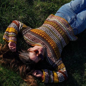 May include: A colorful crochet sweater with a geometric pattern in shades of brown, yellow, and white. The long-sleeved sweater is paired with blue jeans. The person is wearing sunglasses and lying on green grass.