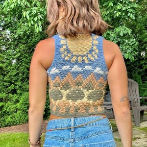 May include: A blue, yellow, and tan crochet top with a geometric design. The sleeveless top features a sunburst pattern around the neckline and a repeating triangle pattern on the front. The top is paired with blue jeans.