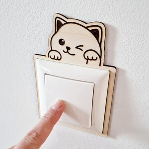 Könnte beinhalten: Eine Holz-Lichtschalterabdeckung mit einem zwinkernden Cartoon-Katzen-Design. Die Abdeckung besteht aus hellem Holz und ist an einer weißen Wand montiert, wobei ein Finger den Schalter drückt.