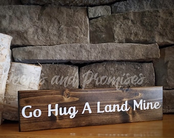May include: A dark brown wooden sign with white text that reads "Go Hug A Land Mine". The sign is sitting on a wooden surface in front of a stone wall.