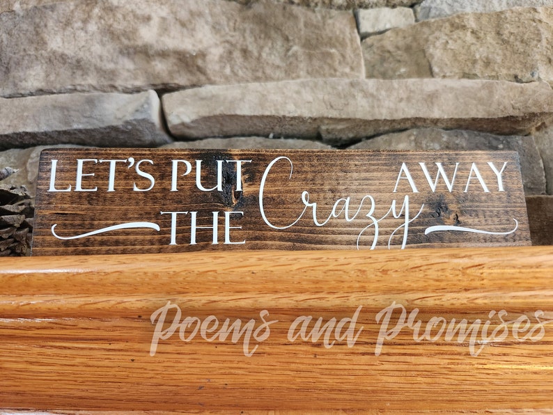May include: A wooden sign with the text "Let's Put The Crazy Away" in white lettering on a dark brown background.