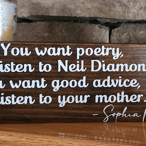May include: A wooden sign with a dark brown stain features a white inscription that reads: "You want poetry, you listen to Neil Diamond. You want good advice, you listen to your mother." The inscription is attributed to Sophia Petrillo.