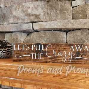 May include: A wooden sign with the text "Let's Put the Crazy Away" in white lettering on a dark brown background. The sign is sitting on a wooden surface.