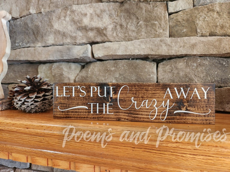 May include: A wooden sign with the text "Let's Put the Crazy Away" in white lettering on a dark brown background. The sign is sitting on a wooden surface.