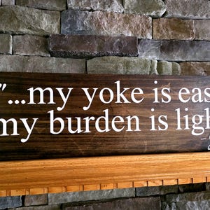 May include: Wooden sign with the quote "...my yoke is easy; my burden is light." in white lettering. The sign is stained dark brown and rests on a wooden shelf in front of a stone wall. The signature "Jesus" is at the bottom.