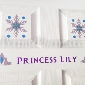 K&ouml;nnte beinhalten: Wei&szlig;e T&uuml;r mit einer lila Aufschrift, die "Princess Lily" lautet. Zwei blaue und lila Schneeflocken-Aufkleber befinden sich &uuml;ber der Aufschrift. Zwei blaue und lila stilisierte Kronen-Aufkleber befinden sich auf beiden Seiten der Aufschrift.