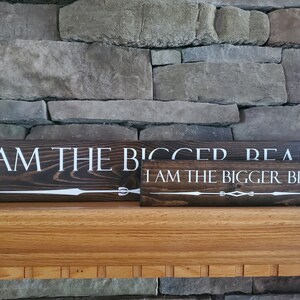 May include: Two wooden signs with white text on a dark brown background. The top sign reads "I am the bigger read". The bottom sign reads "I am the bigger bear".