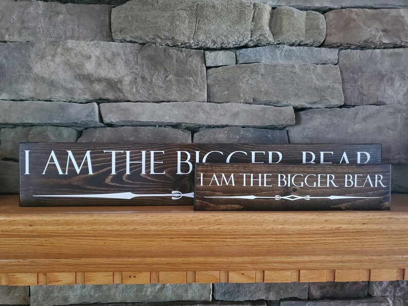 May include: Two wooden signs with white text on a dark brown background. The top sign reads "I am the bigger read". The bottom sign reads "I am the bigger bear".