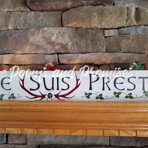 May include: White wooden sign with the text "Je Suis Prest" in black lettering. The sign features a red antler design with a white flower in the center. Green vines are painted on either side of the sign.