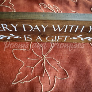 May include: Wooden sign with the text "Every day with you is a gift" in white lettering. The sign has a rustic brown finish and is decorated with small white leaf sprigs.