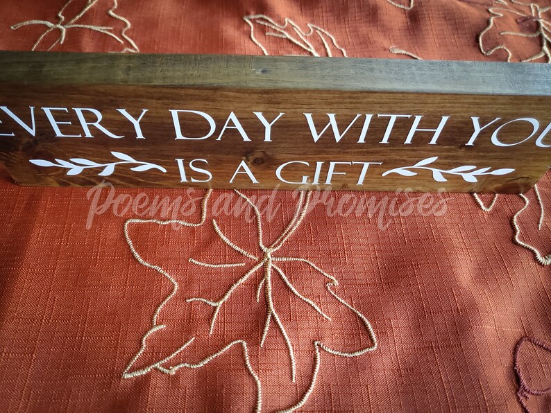 May include: Wooden sign with the text "Every day with you is a gift" in white lettering. The sign has a rustic brown finish and is decorated with small white leaf sprigs.