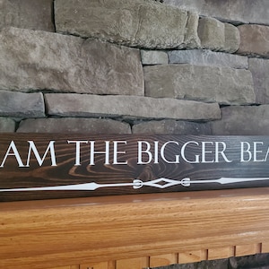 May include: A dark brown wooden sign with white lettering that reads "I AM THE BIGGER BEAR". The sign has a decorative white design at the bottom.