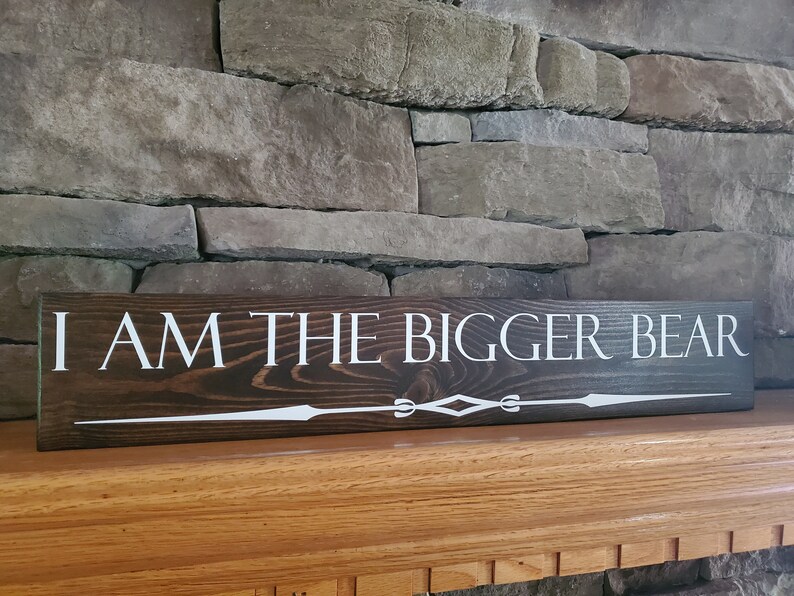 May include: A dark brown wooden sign with white lettering that reads "I AM THE BIGGER BEAR". The sign has a decorative white design at the bottom.