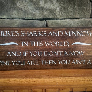 May include: A wooden sign with a dark brown stain and white lettering. The sign reads "THERE'S SHARKS AND MINNOWS IN THIS WORLD, AND IF YOU DON'T KNOW WHICH ONE YOU ARE, THEN YOU AIN'T A SHARK".