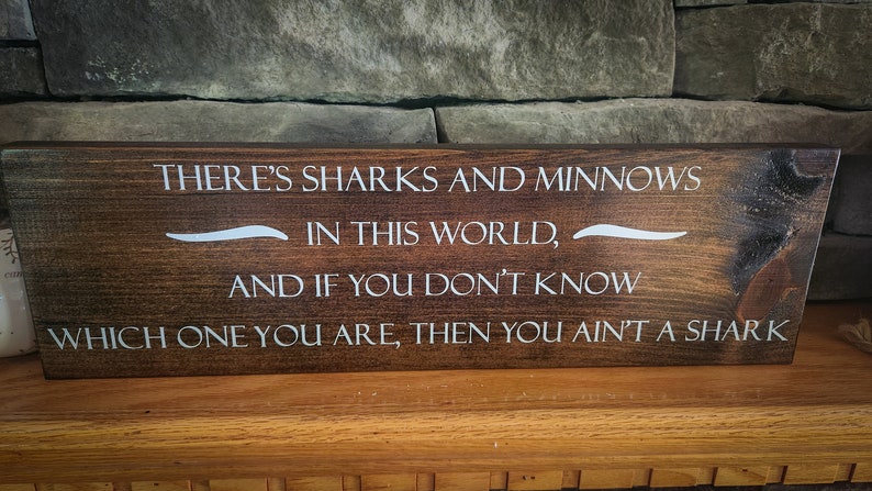 May include: A wooden sign with a dark brown stain and white lettering. The sign reads "THERE'S SHARKS AND MINNOWS IN THIS WORLD, AND IF YOU DON'T KNOW WHICH ONE YOU ARE, THEN YOU AIN'T A SHARK".