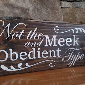 Könnte beinhalten: Ein Holzschild mit dem Text "Not the Meek and Obedient Type" in weißen Buchstaben auf einem dunkelbraunen Hintergrund.