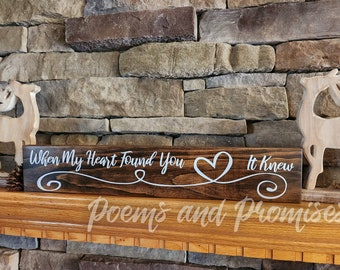May include: A dark brown wooden sign with white script reading "When My Heart Found You It Knew" and a heart symbol. The sign, with the words "Poems and Promises", is displayed on a wooden mantelpiece, against a stone wall.