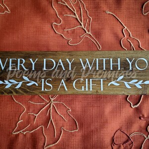 May include: A wooden sign with the text "EVERY DAY WITH YOU IS A GIFT" in white lettering. The sign has a brown wood grain and is decorated with white leafy vines.