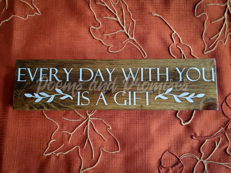 May include: A wooden sign with the text "EVERY DAY WITH YOU IS A GIFT" in white lettering. The sign has a brown wood grain and is decorated with white leafy vines.