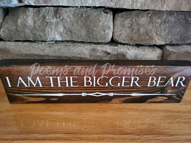 May include: A dark brown wooden sign with the text "I AM THE BIGGER BEAR" in white lettering. The sign has a decorative design at the bottom.