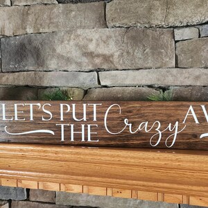 May include: A brown wooden sign with white lettering that reads "Let's Put The Crazy Away". The sign is sitting on a wooden mantel above a brick fireplace.