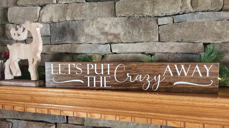 May include: A brown wooden sign with white lettering that reads "Let's Put The Crazy Away". The sign is sitting on a wooden mantel above a brick fireplace.