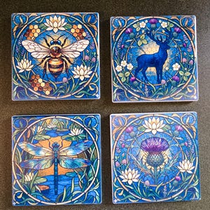 Puede incluir: Juego de cuatro posavasos cuadrados con diseños Art Nouveau. Cada posavasos presenta una imagen diferente: una abeja, un ciervo, una libélula y un cardo. Los diseños están sobre un fondo azul con patrones florales y geométricos.