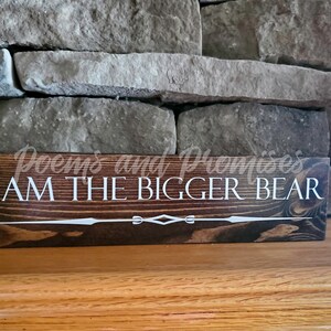 May include: A dark brown wooden sign with white text that reads "Poems and Promises I AM THE BIGGER BEAR". The sign has a decorative white design at the bottom.