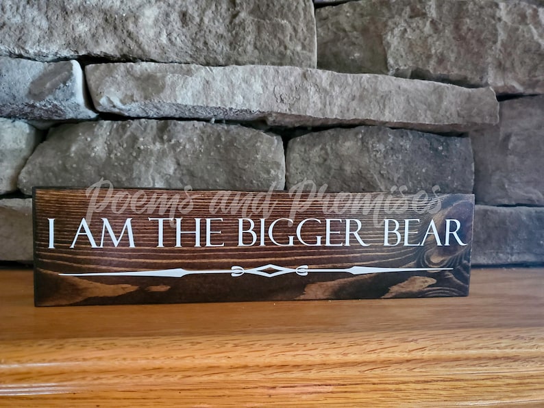 May include: A dark brown wooden sign with white text that reads "Poems and Promises I AM THE BIGGER BEAR". The sign has a decorative white design at the bottom.