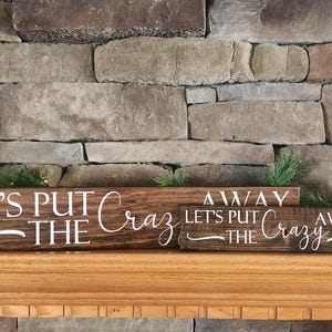 May include: Three wooden signs with the text "Let's Put The Crazy Away" on a brown brick fireplace mantel. The signs are stacked on top of each other.