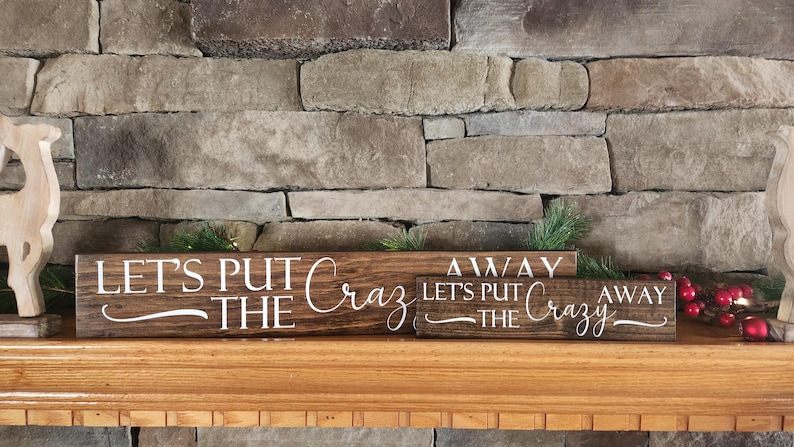 May include: Three wooden signs with the text "Let's Put The Crazy Away" on a brown brick fireplace mantel. The signs are stacked on top of each other.