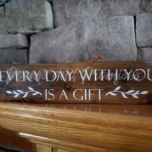 May include: A wooden sign with the text "EVERYDAY WITH YOU IS A GIFT" in white lettering. The sign has a brown stain and is decorated with white leafy vines.