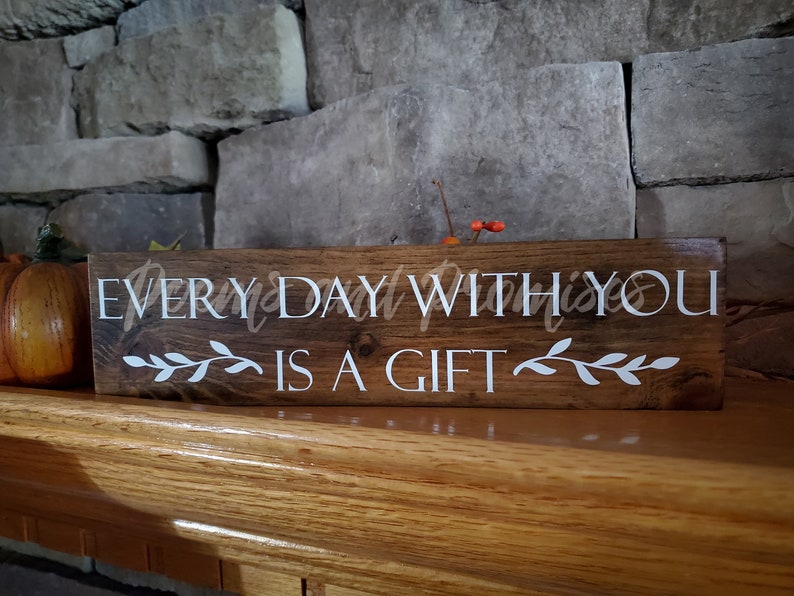 May include: A wooden sign with the text "EVERYDAY WITH YOU IS A GIFT" in white lettering. The sign has a brown stain and is decorated with white leafy vines.
