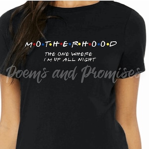 May include: Black t-shirt with the word "MOTHERHOOD" in white, with the "O"s in red, yellow, blue, and orange. Below, the text reads "THE ONE WHERE I'M UP ALL NIGHT". The shirt is a crew neck.