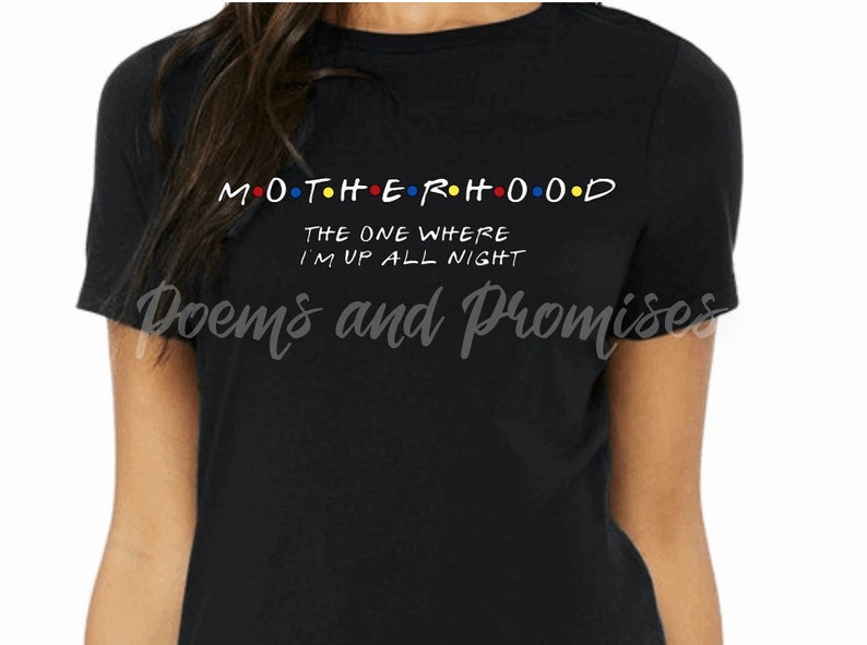 May include: Black t-shirt with the word "MOTHERHOOD" in white, with the "O"s in red, yellow, blue, and orange. Below, the text reads "THE ONE WHERE I'M UP ALL NIGHT". The shirt is a crew neck.