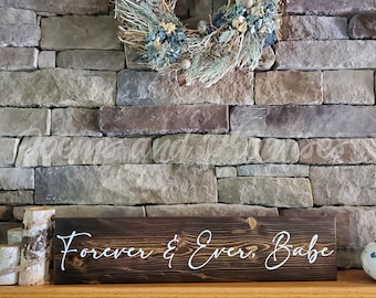 May include: A dark brown wooden sign with the text "Forever & Ever, Babe" written in white cursive script. The sign is sitting on a wooden mantel above a stone fireplace.