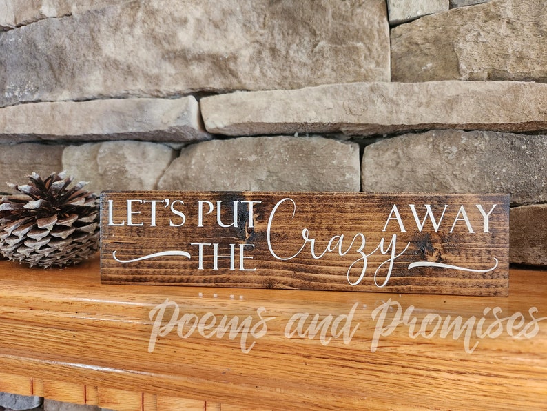 May include: A rustic wooden sign with the text "Let's Put The Crazy Away" in white lettering. The sign has a dark brown stain and a distressed look.