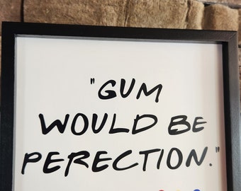 Op de afbeelding: Een ingelijst print met de quote "Gum would be perfection." - Chandler. De quote staat in zwarte tekst op een witte achtergrond. Er zijn zes gekleurde stippen onder de quote.