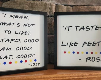 Op de afbeelding: Twee ingelijste prints met zwarte lijsten en witte achtergronden. De linker print heeft de tekst "Ik bedoel, wat is er niet om van te houden? Pudding, lekker. Jam, lekker. Vlees, lekker!" met een rij rode, gele en blauwe stippen eronder. De rechter print heeft de tekst "Het smaakt naar voeten!" met een rij rode, gele en blauwe stippen eronder en de tekst "Ross" onder de stippen.