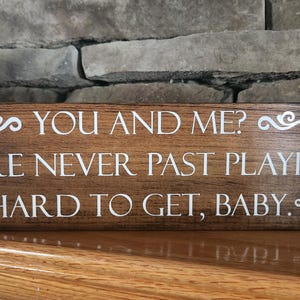 May include: A wooden sign with the text "YOU AND ME? WE'RE NEVER PAST PLAYING HARD TO GET, BABY." in white lettering.