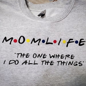 以下が含まれることがあります： グレーのトレーナーに、黒い文字で「M.O.M.L.I.F.E. "The One Where I Do All The Things"」と書かれている