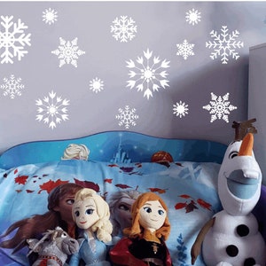 May include: A blue and white bed with a white headboard and a blue and white comforter. There are several white snowflake decals on the wall behind the bed. There are four dolls sitting on the bed, and a large Olaf plush toy sitting on the bed.