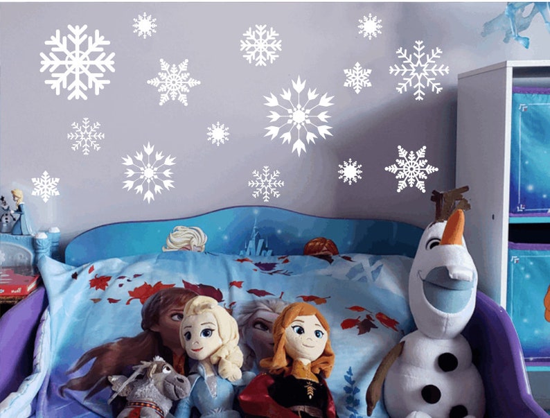 May include: A blue and white bed with a white headboard and a blue and white comforter. There are several white snowflake decals on the wall behind the bed. There are four dolls sitting on the bed, and a large Olaf plush toy sitting on the bed.