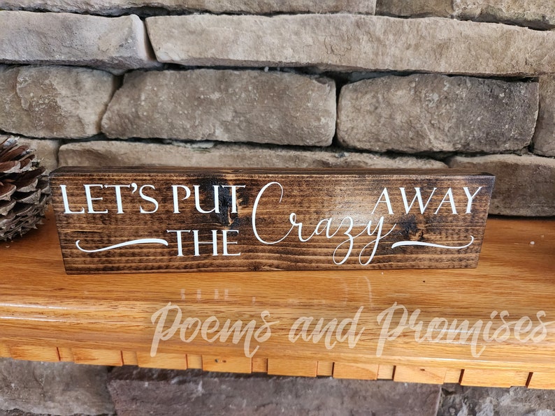 May include: A wooden sign with the text "Let's Put The Crazy Away" in white cursive lettering on a dark brown background.