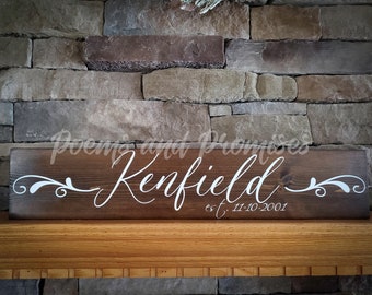 May include: A rectangular, dark brown wooden sign with the name "Kenfield" in white script. Below the name is "est. 11-10-2001". The sign has white decorative flourishes on each side. The sign is displayed on a wooden mantle.