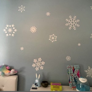 May include: A light blue wall with white snowflake decals. There are several snowflakes of varying sizes and designs. The wall is decorated with a stuffed animal, a toy, and a humidifier.