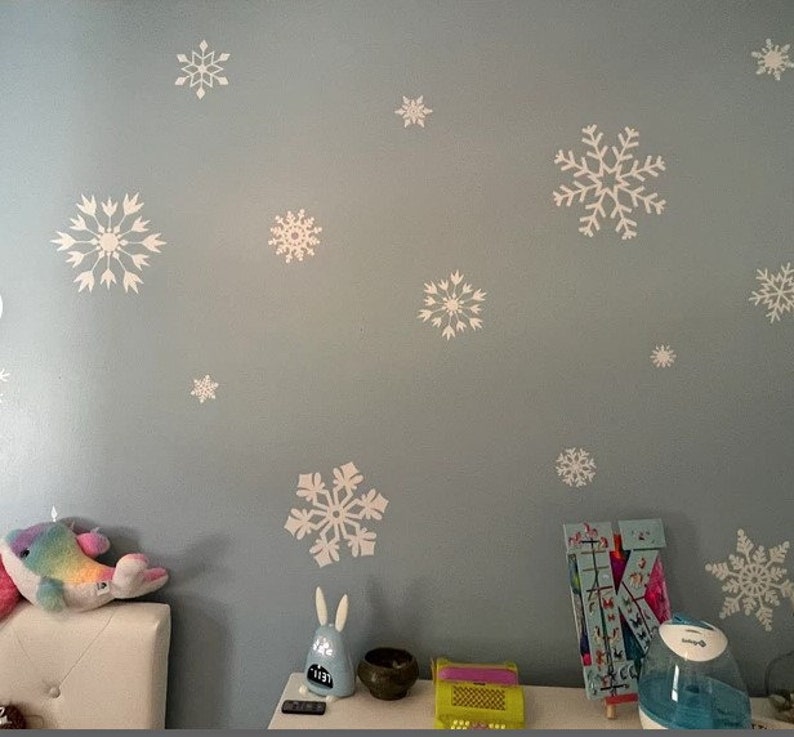 May include: A light blue wall with white snowflake decals. There are several snowflakes of varying sizes and designs. The wall is decorated with a stuffed animal, a toy, and a humidifier.
